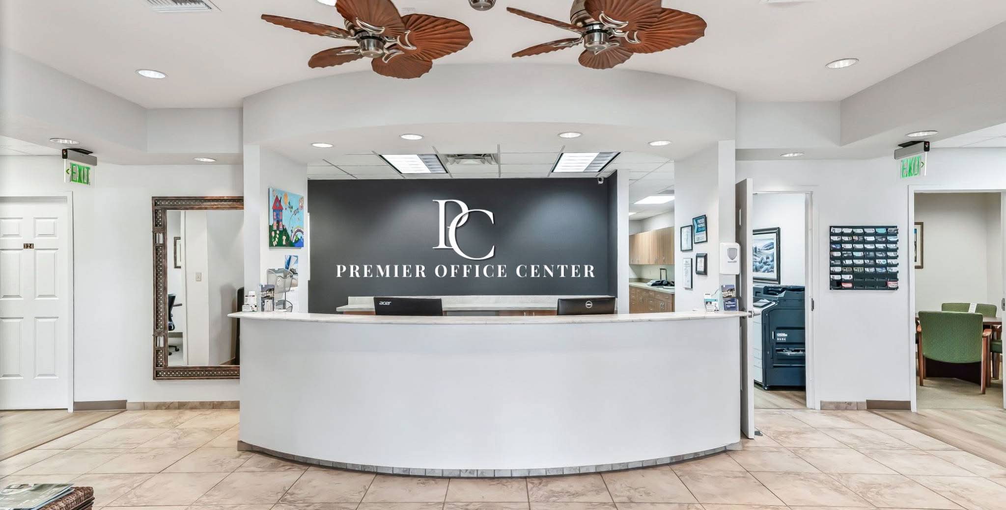 A modern office lobby featuring a curved white reception desk with "Premier Office Center" on a dark wall behind. The room is bright with two ceiling fans.