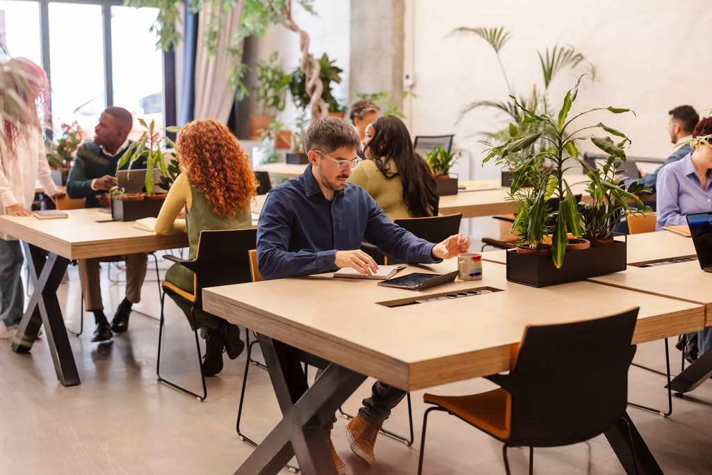 A diverse group of professionals working in a modern, plant-filled coworking space in Austin, TX, with a man in the foreground using a laptop and notebook.