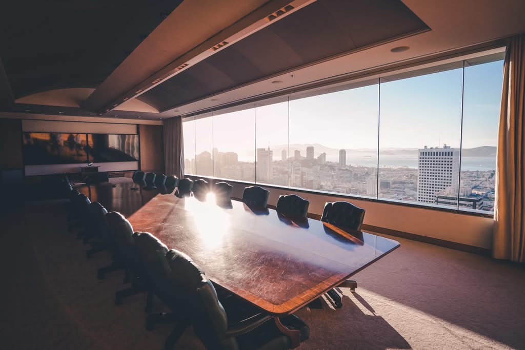 A premium financial advisor meeting room featuring a long polished wood conference table, high-back leather chairs, and a panoramic wall window showing the bright San Francisco city skyline and bay area at sunset.