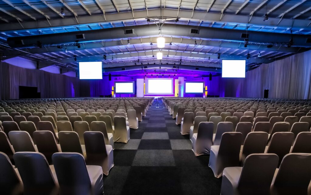Thousands of empty chairs with white seat covers arranged in precise rows facing a large stage with blank projection screens under blue and purple ambient lighting at a training room rental facility in Wildwood, Florida.