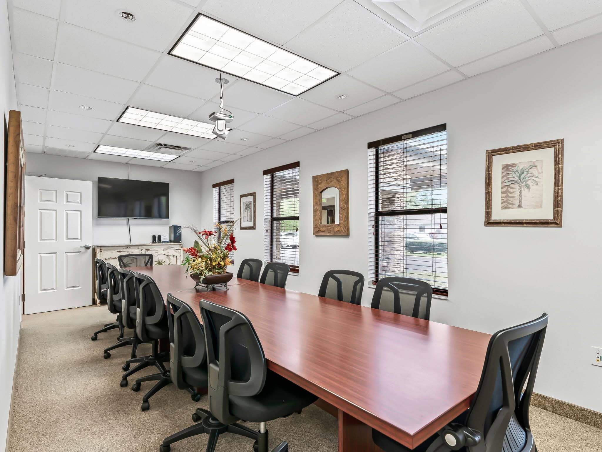 A spacious conference room rental in Wildwood, FL featuring a long wood boardroom table, ergonomic mesh chairs, wall-mounted display screen, and natural light.