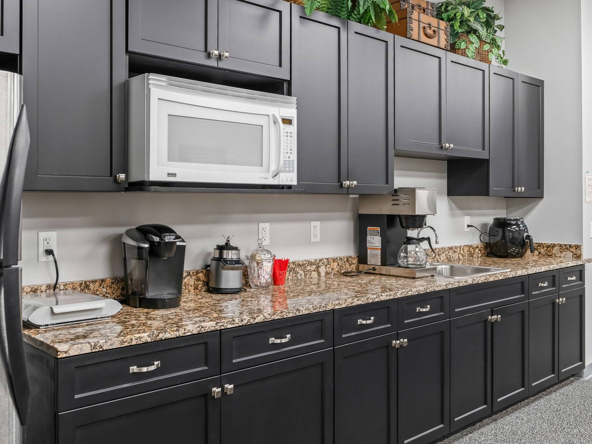 A modern office breakroom kitchen in Ocala, FL featuring dark cabinetry, granite countertops, coffee makers, microwave, and small appliances.