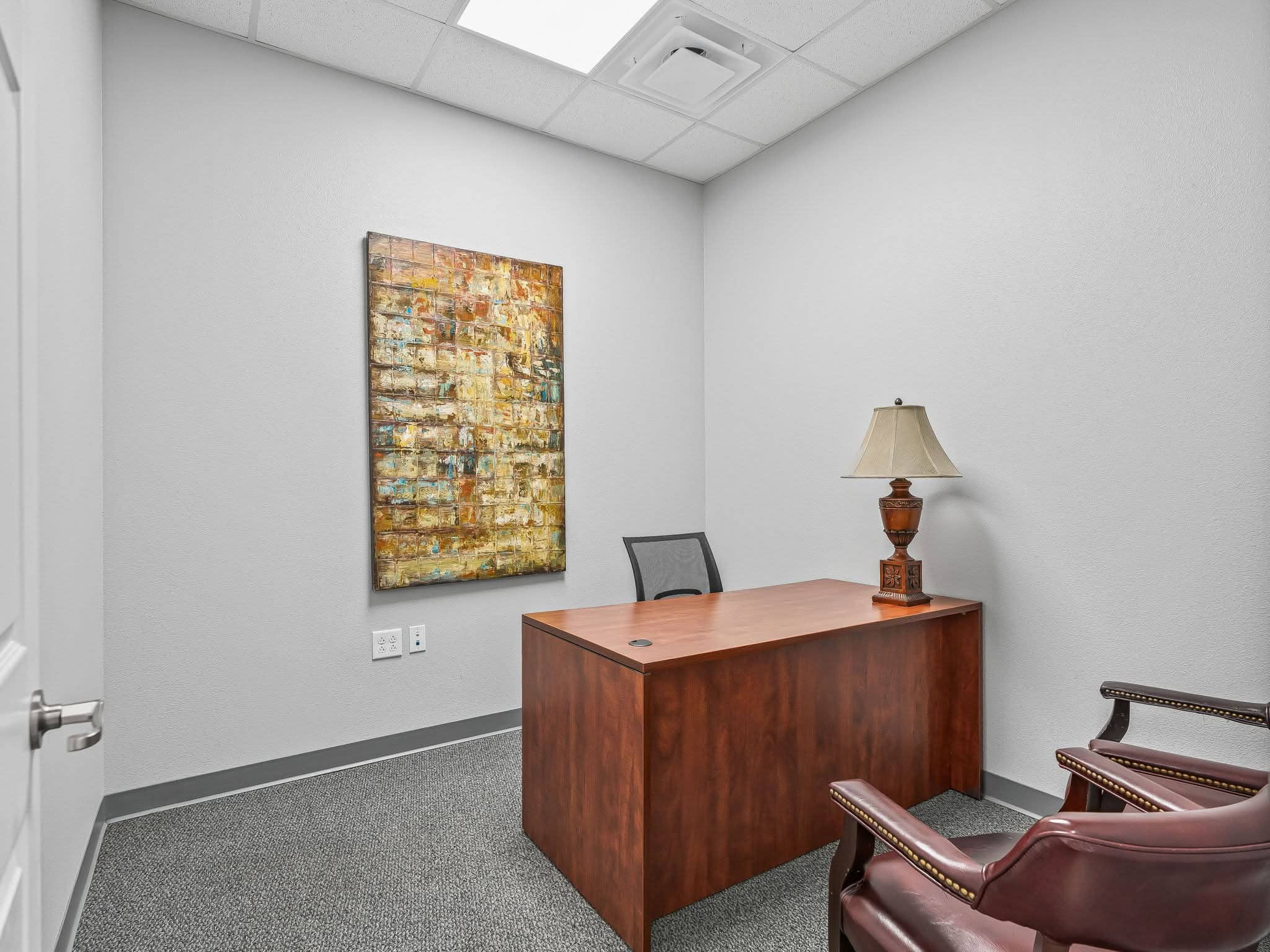A fully furnished private executive suite in Mount Dora, FL featuring a warm wood desk, lamp, executive seating, and abstract wall art.