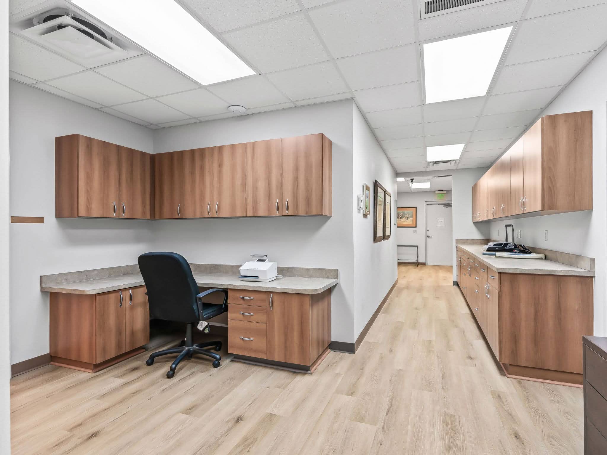 A modern office breakroom and administrative mail handling work area in Mount Dora, FL featuring light wood cabinetry, vinyl plank flooring, built-in desks, and office supplies.