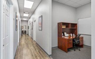 A modern office hallway with gray walls and wood flooring with amenities. On the right, there's a wooden desk with a black chair and a whiteboard, creating a clean, professional atmosphere.