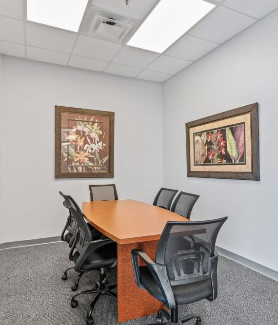 Small meeting room with a wooden table and six black mesh chairs. Two floral paintings adorn the gray walls, creating a calm atmosphere.