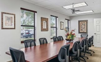A long conference room with a wooden table, black chairs, and a floral centerpiece. Windows line one wall, and framed art decorates the light gray walls.