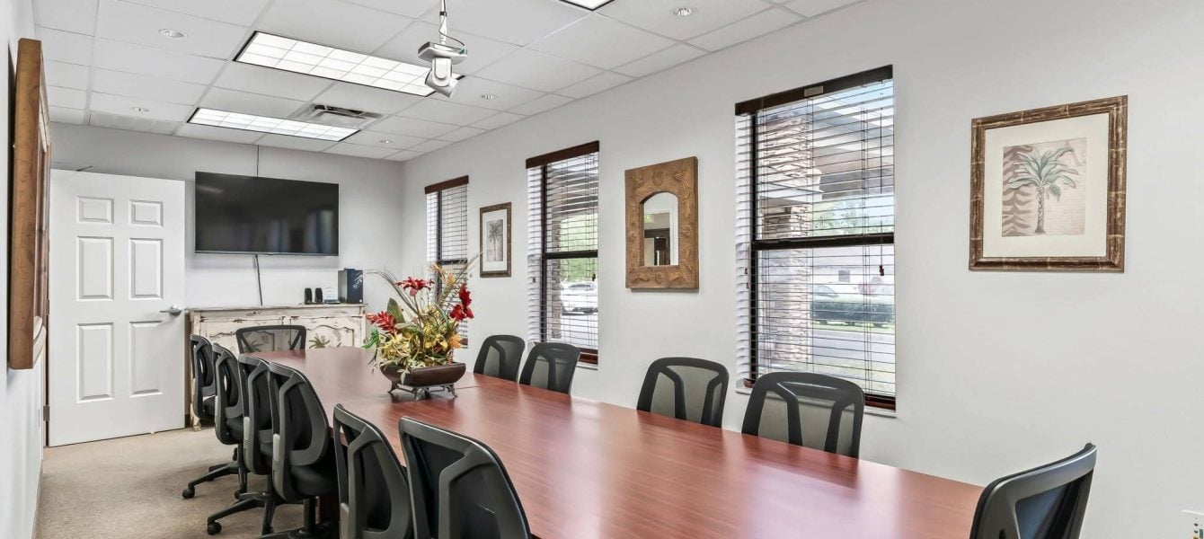 Large executive conference room featuring a long wooden conference table, ergonomic black mesh chairs, a wall-mounted TV screen, and large windows in The Villages, FL.