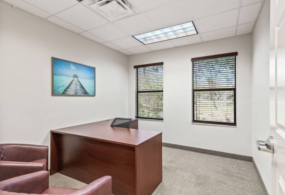 An office space for custom shed consultation in Ocala, FL featuring a large mahogany L-shaped desk, leather client armchairs, framed tropical dock artwork, and two windows with blinds.