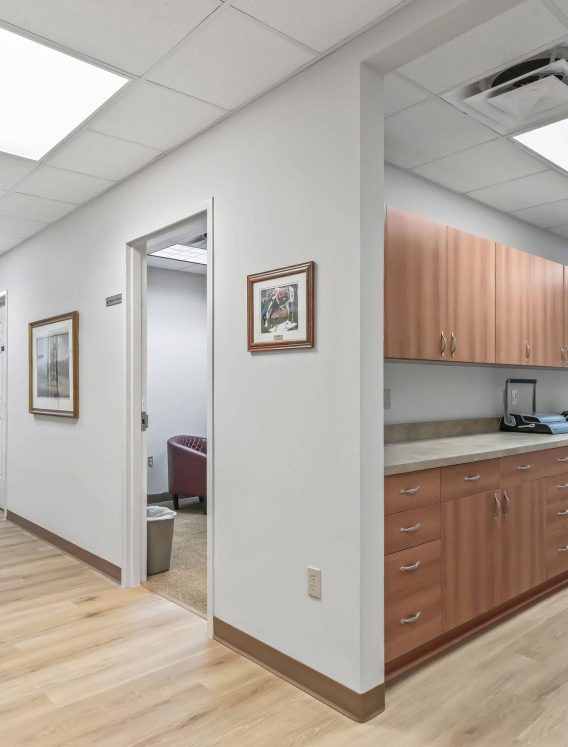 A modern executive suites hallway in Mount Dora, FL with light wood flooring, framed wall art, an open private office doorway, and an administrative work area featuring built-in cabinets, mail equipment, and office printing machinery.