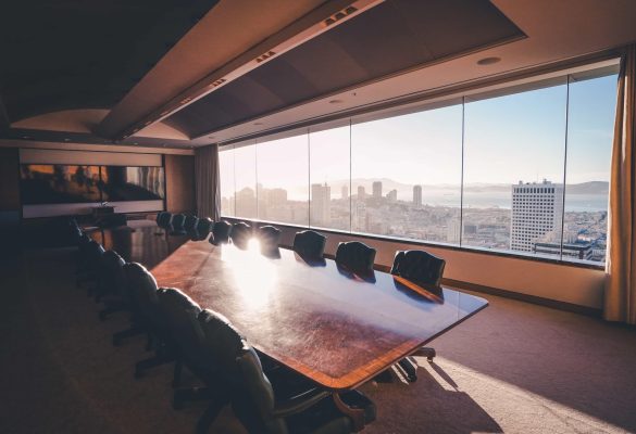 A premium financial advisor meeting room featuring a long polished wood conference table, high-back leather chairs, and a panoramic wall window showing the bright San Francisco city skyline and bay area at sunset.