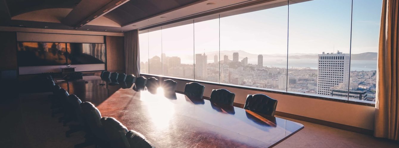 A premium financial advisor meeting room featuring a long polished wood conference table, high-back leather chairs, and a panoramic wall window showing the bright San Francisco city skyline and bay area at sunset.