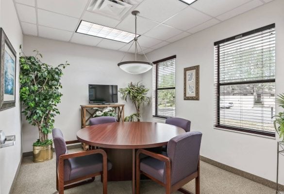 Bright private meeting room featuring a round wooden table, four purple armchairs, potted indoor plants, large windows, and a central pendant light in Ocala, FL.