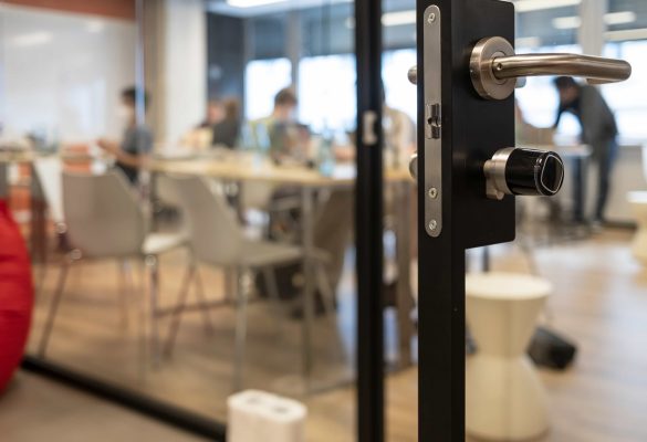 A close-up of a secure black glass door handle with a modern lock mechanism, looking into a blurred conference room where a business meeting is taking place in a HIPAA-compliant office space in Denver, Colorado.