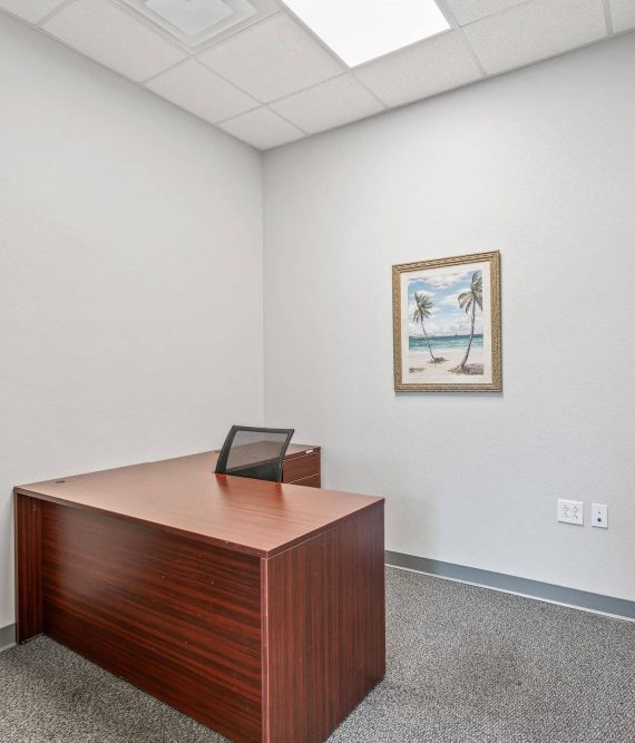 A private office space in Ocala, FL featuring an L-shaped wooden executive desk, an ergonomic task chair, a leather armchair, carpet flooring, and beach artwork on a light wall.