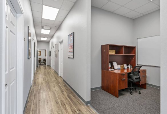 A bright office hallway with wood-look flooring leading past doors to a private workstation niche with a wooden desk and chair in a professional office suite in Ocala, FL.