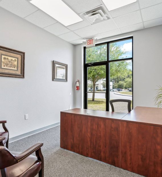 A modern sales office in Ocala, FL featuring a large wooden desk, leather armchairs, potted plant, framed wall art, and a glass exit door revealing green trees outside.