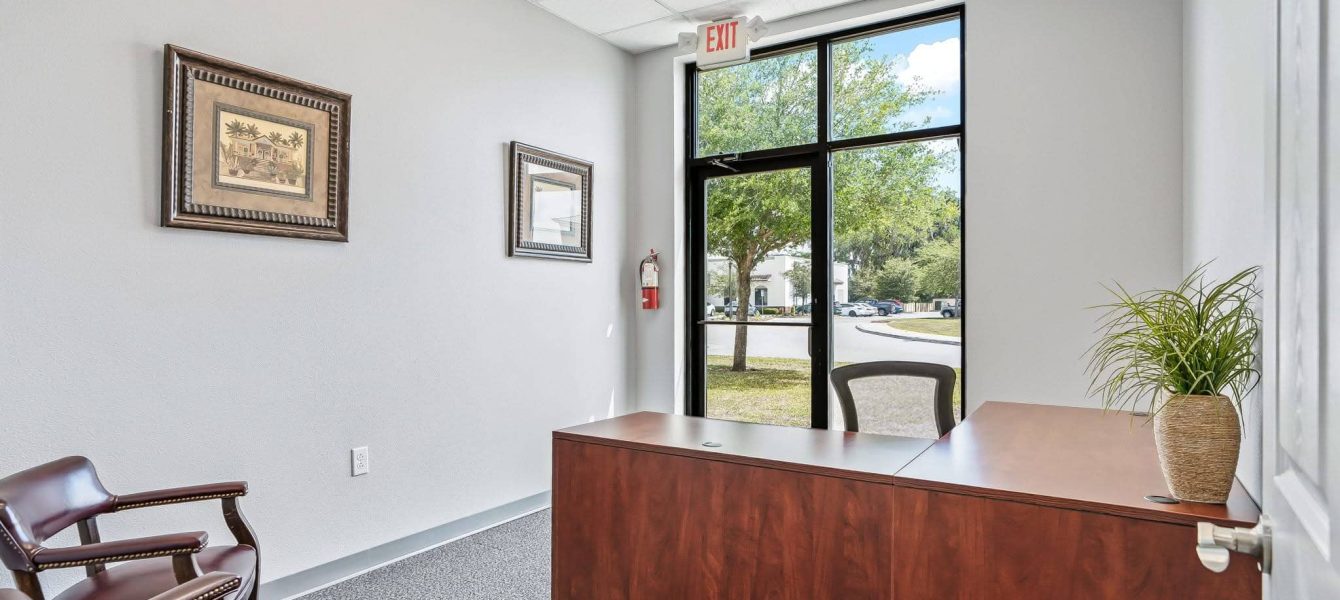 A modern sales office in Ocala, FL featuring a large wooden desk, leather armchairs, potted plant, framed wall art, and a glass exit door revealing green trees outside.
