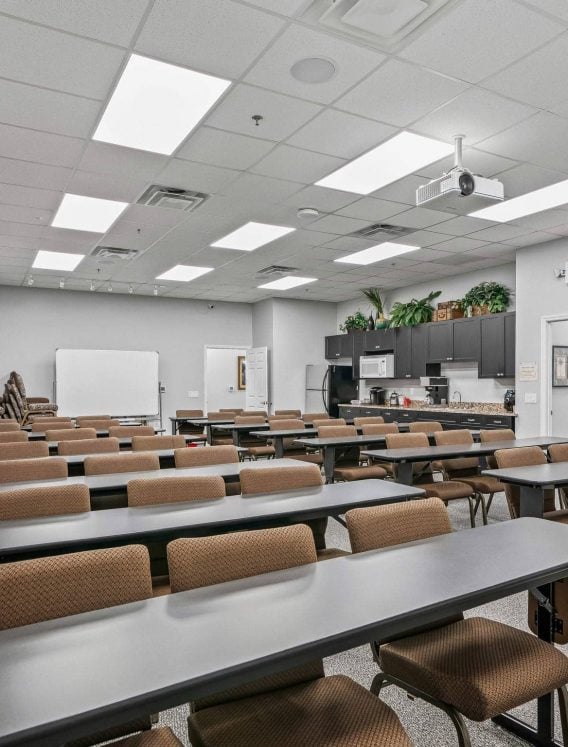 Large classroom-style corporate training room rentals with rows of grey tables, brown padded chairs, ceiling projector, whiteboard, and kitchenette in The Villages, FL.