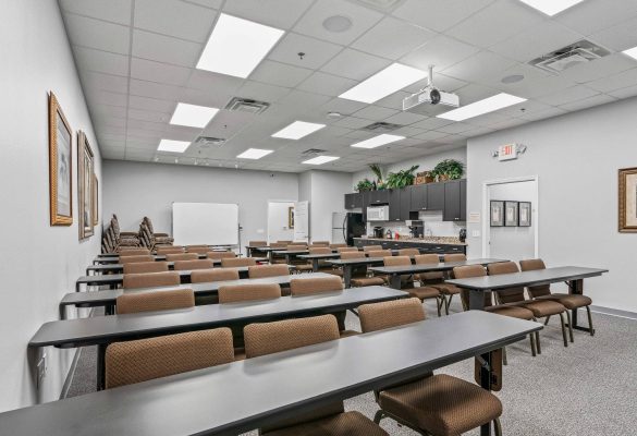 Large classroom-style corporate training room rentals with rows of grey tables, brown padded chairs, ceiling projector, whiteboard, and kitchenette in The Villages, FL.